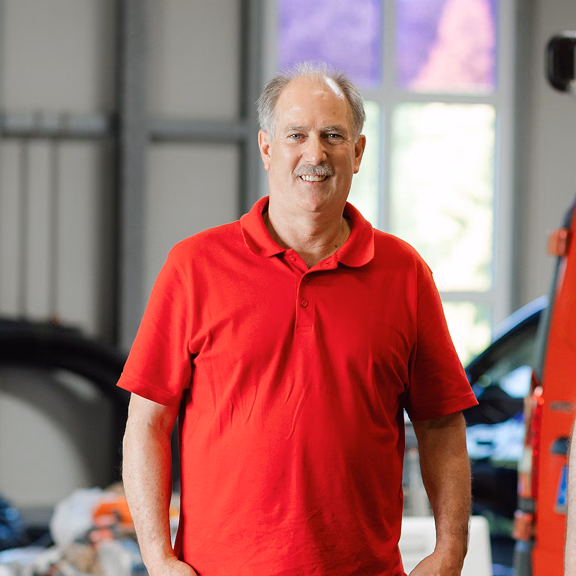Lächelnder älterer Mann mit grauen Haaren im roten Poloshirt steht in einer Werkstatthalle